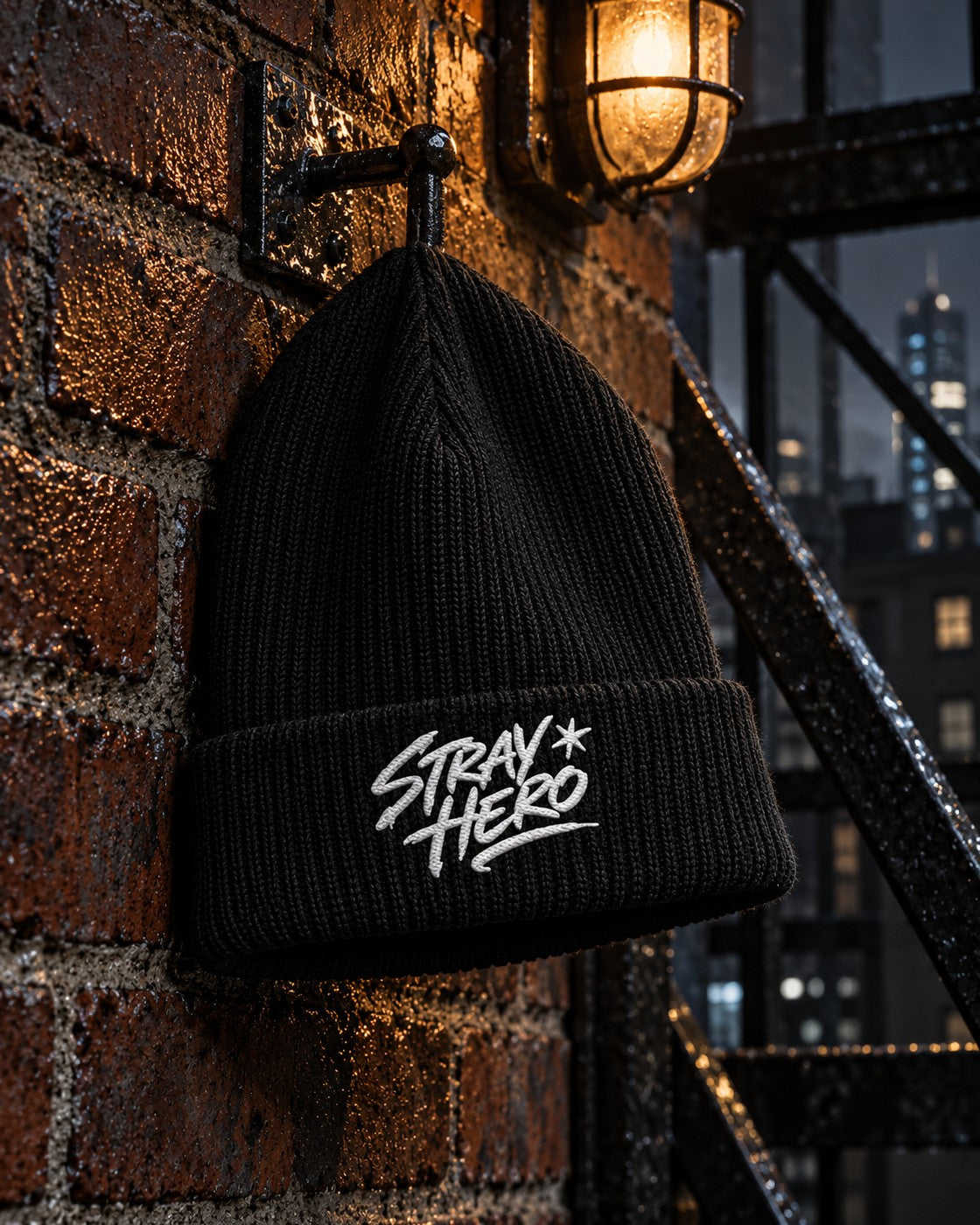 Signature Fisherman Beanie, front embroidery, black, hanging from a hook on a brick wall beside a fire escape at night — AI-generated visual