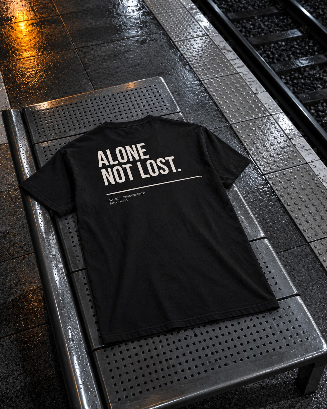 Alone Not Lost Tee Typography, back print, black, laid on a perforated metal bench on a train platform at night — AI-generated visual