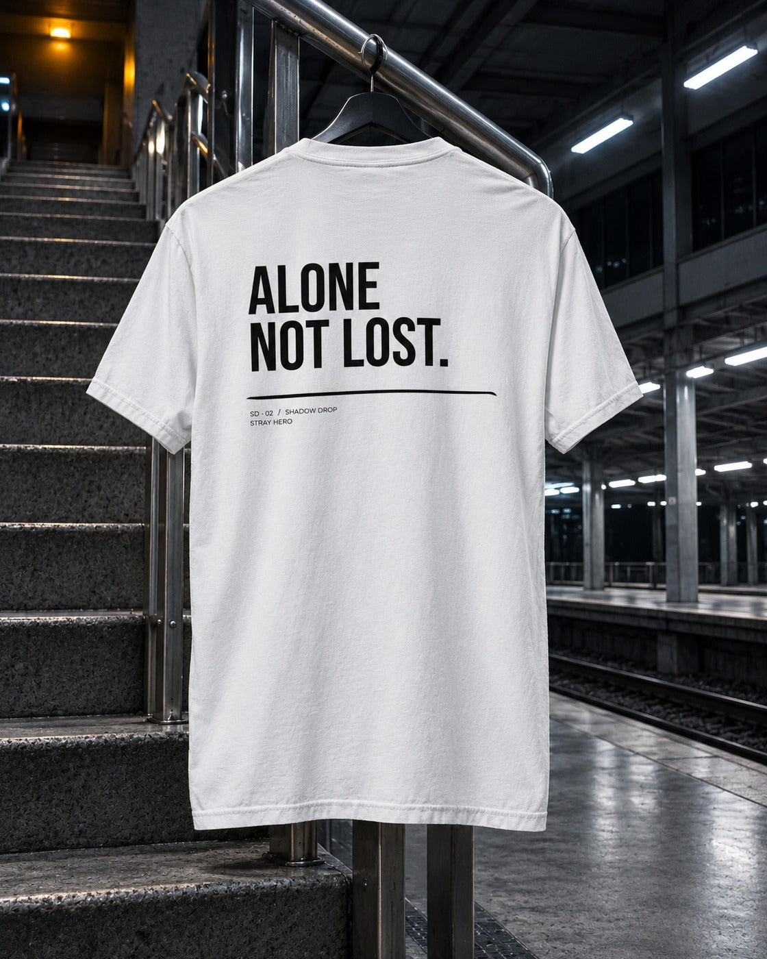 Alone Not Lost Tee White, back print, white, hanging on a stair railing on a train platform at night — AI-generated visual