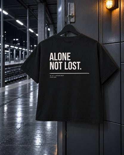 Alone Not Lost Women's Tee, back print, black, hanging on a locker door under a wall lamp on a train platform — AI-generated visual