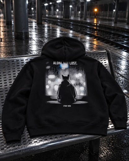 Alone Not Lost Hoodie, back print, black, laid on a metal bench on a wet train platform at night — AI-generated visual