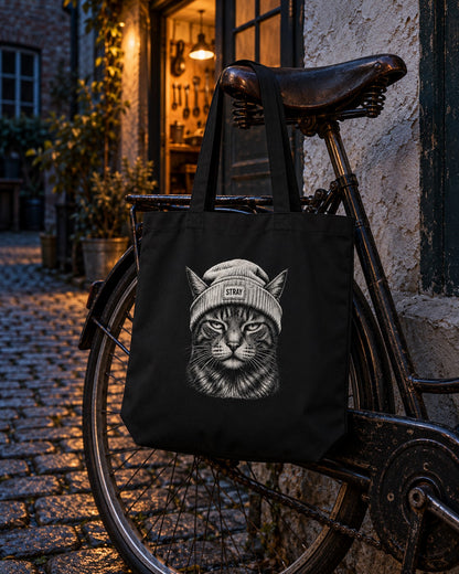 Beanie Cat Organic Tote Ghost, front print, black, hanging from a vintage bicycle in a cobbled lane at night — AI-generated visual