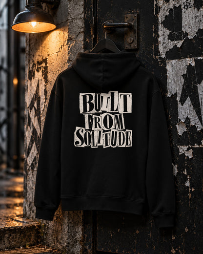 Built From Solitude Hoodie Paste-Up, back print, black, hanging on a weathered pillar under a wall lamp at night — AI-generated visual