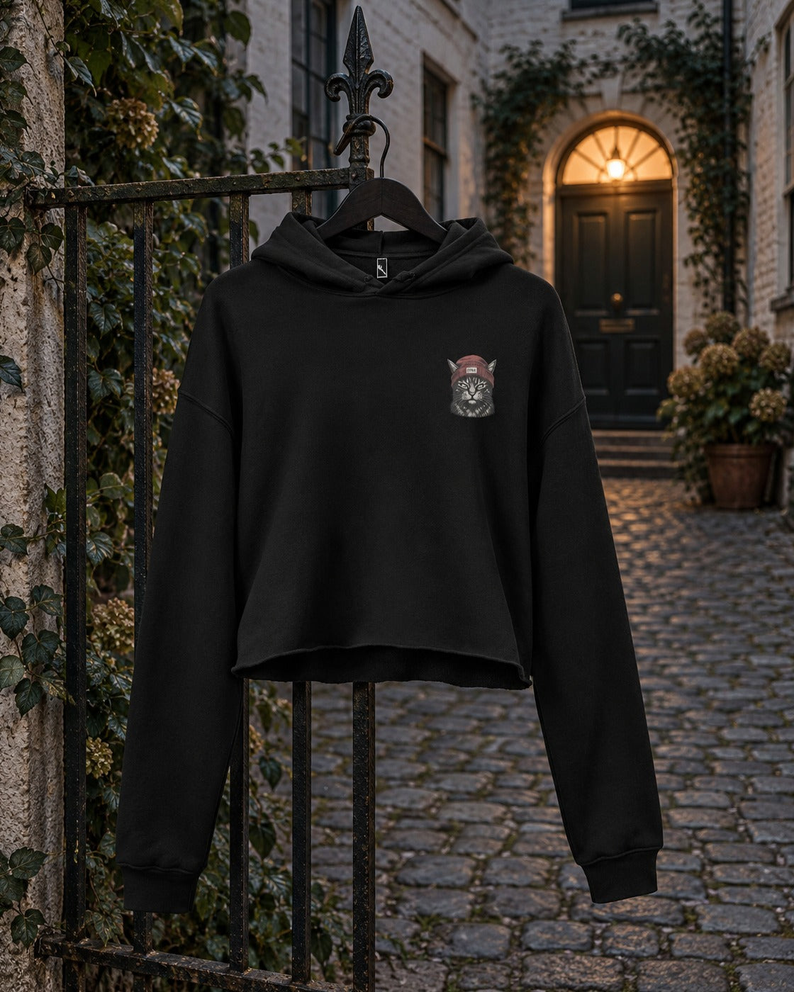 Cat Badge Rogue Crop Hoodie, front print, black, hanging on an iron garden gate in a cobbled courtyard at night — AI-generated visual