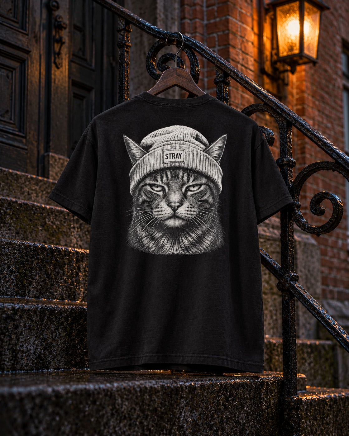 Beanie Cat Tee Ghost, back print, black, hanging on an iron stair railing under a street lamp at night — AI-generated visual
