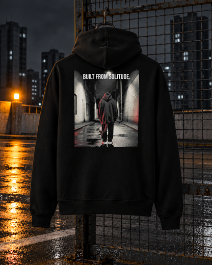 Built From Solitude Hoodie, back print, black, hanging on a fence on a parking deck at night — AI-generated visual