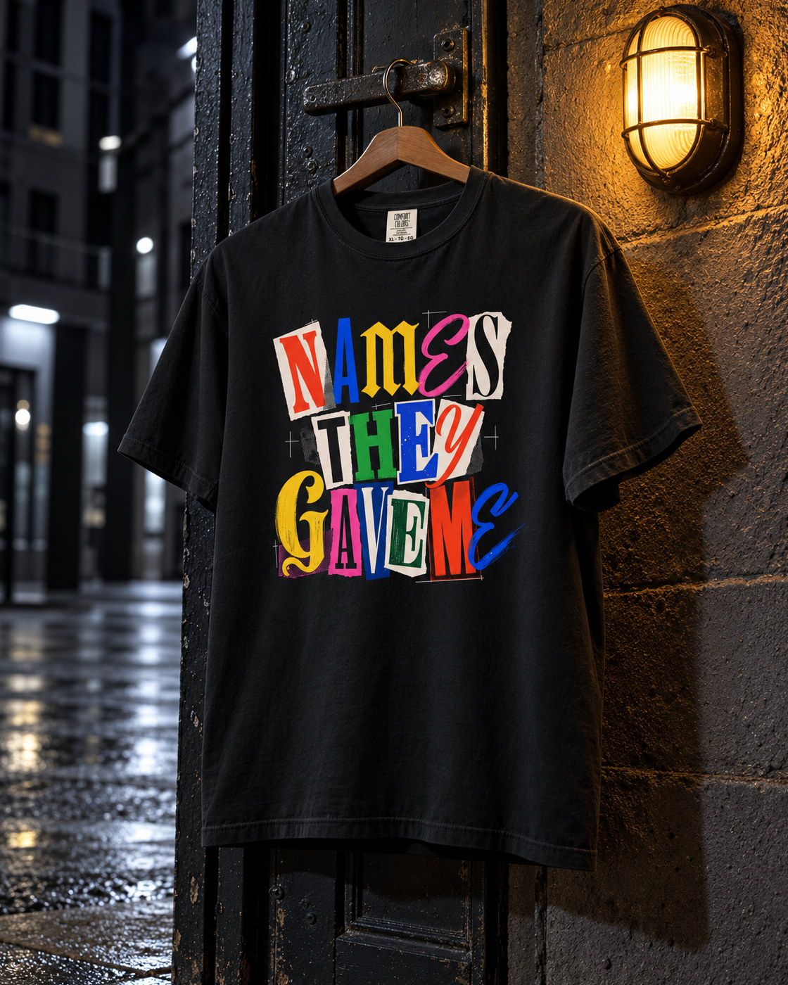 Names They Gave Me Tee, front print, black, hanging under a wall lamp in a backyard at night — AI-generated visual