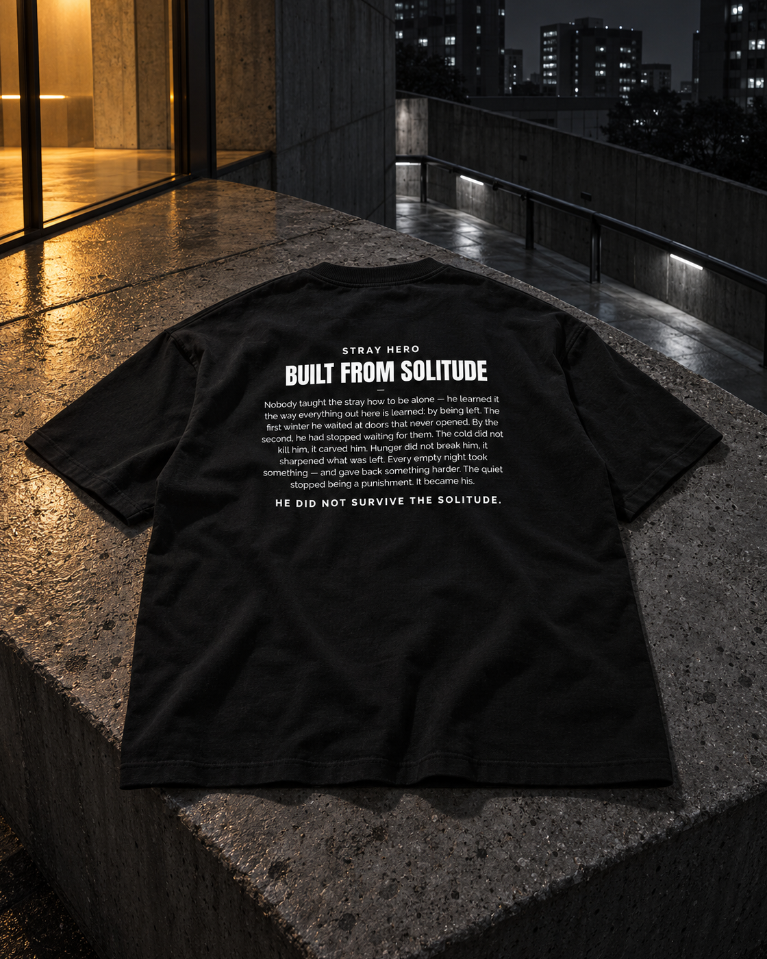 Built From Solitude Story Tee Black, back print, black, laid on a concrete ledge on a parking deck at night — AI-generated visual