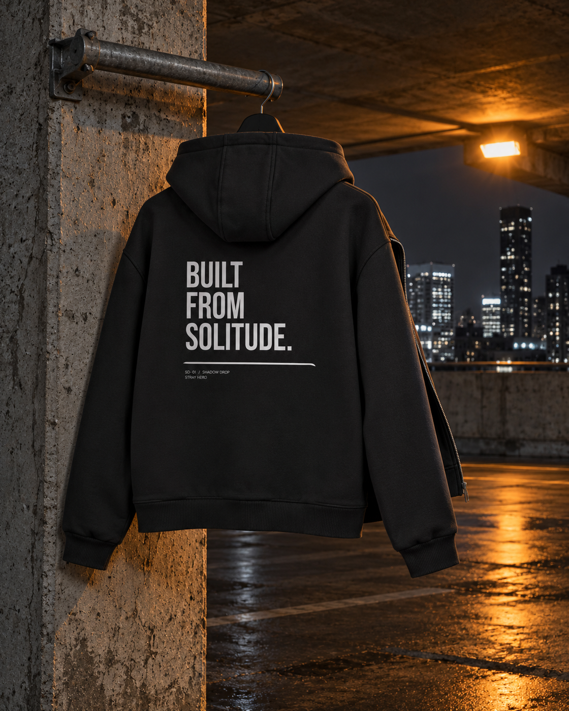 Built From Solitude Zip Hoodie, back print, black, hanging on a concrete pillar under a lamp — AI-generated visual