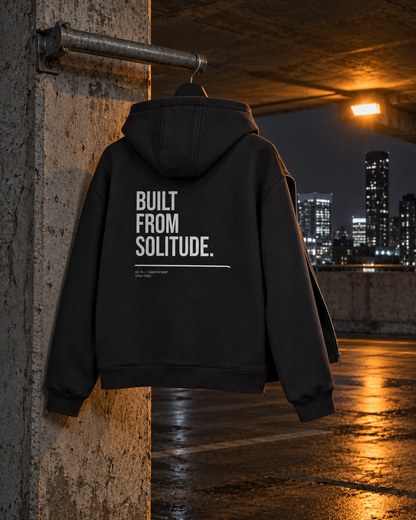 Built From Solitude Zip Hoodie, back print, black, hanging on a concrete pillar under a lamp — AI-generated visual