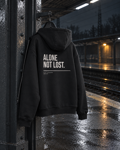 Alone Not Lost Zip Hoodie, back print, black, hanging on a steel pillar on a wet train platform at night — AI-generated visual
