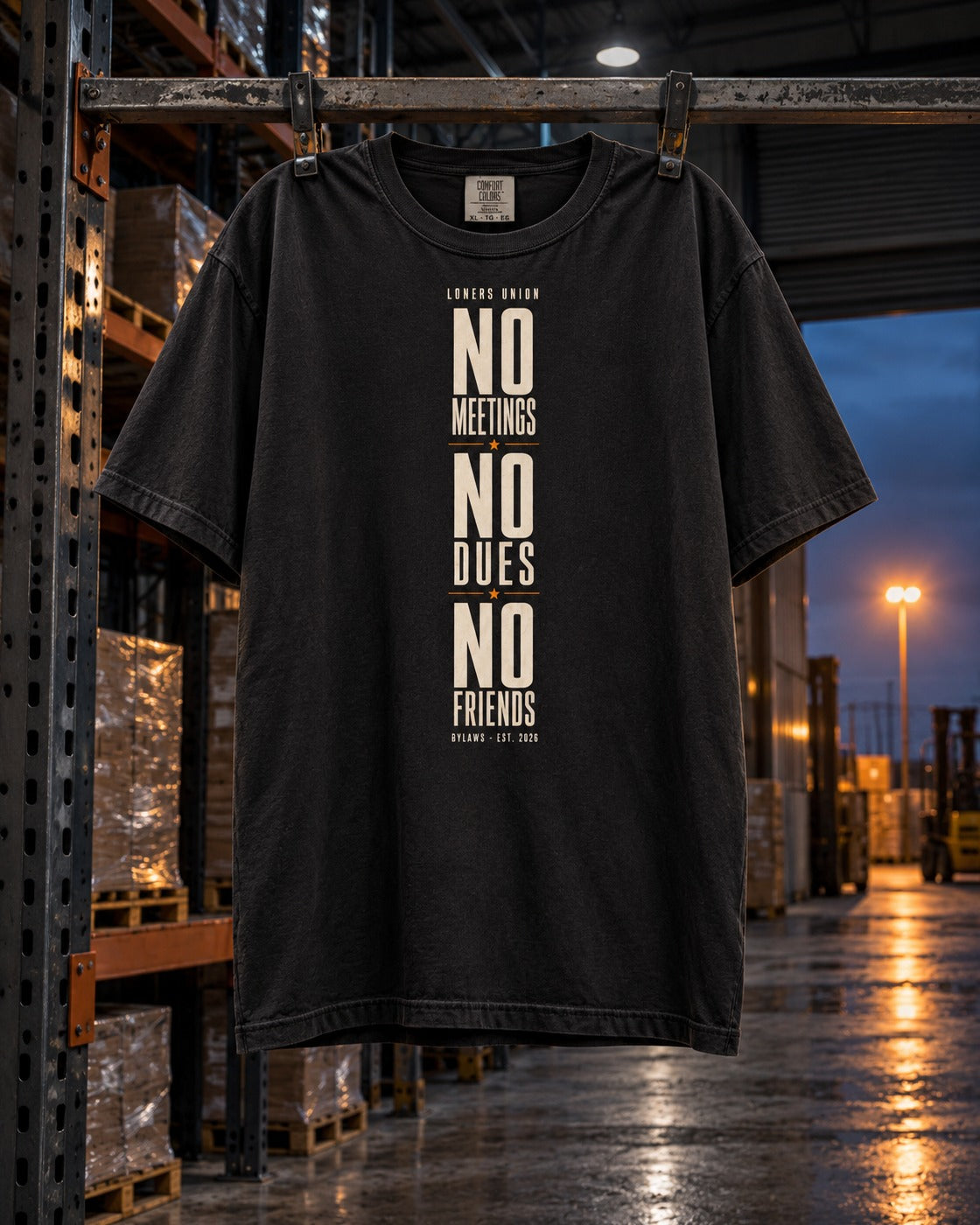 No Meetings No Dues No Friends Tee, front print, black, hanging from a warehouse rack in a wet loading yard at night — AI-generated visual