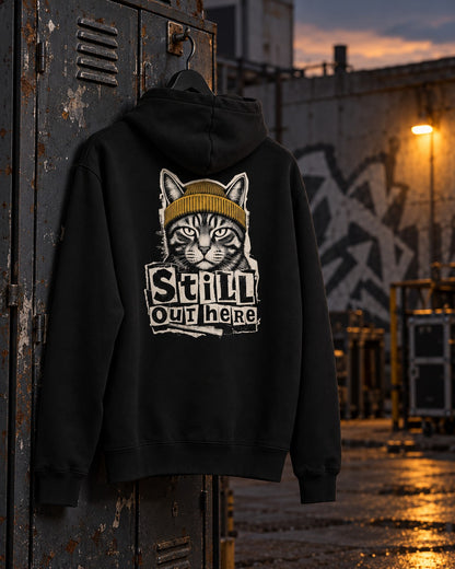 Still Out Here Hoodie, back print, black, hanging on a rusty locker door in a yard at sunset — AI-generated visual