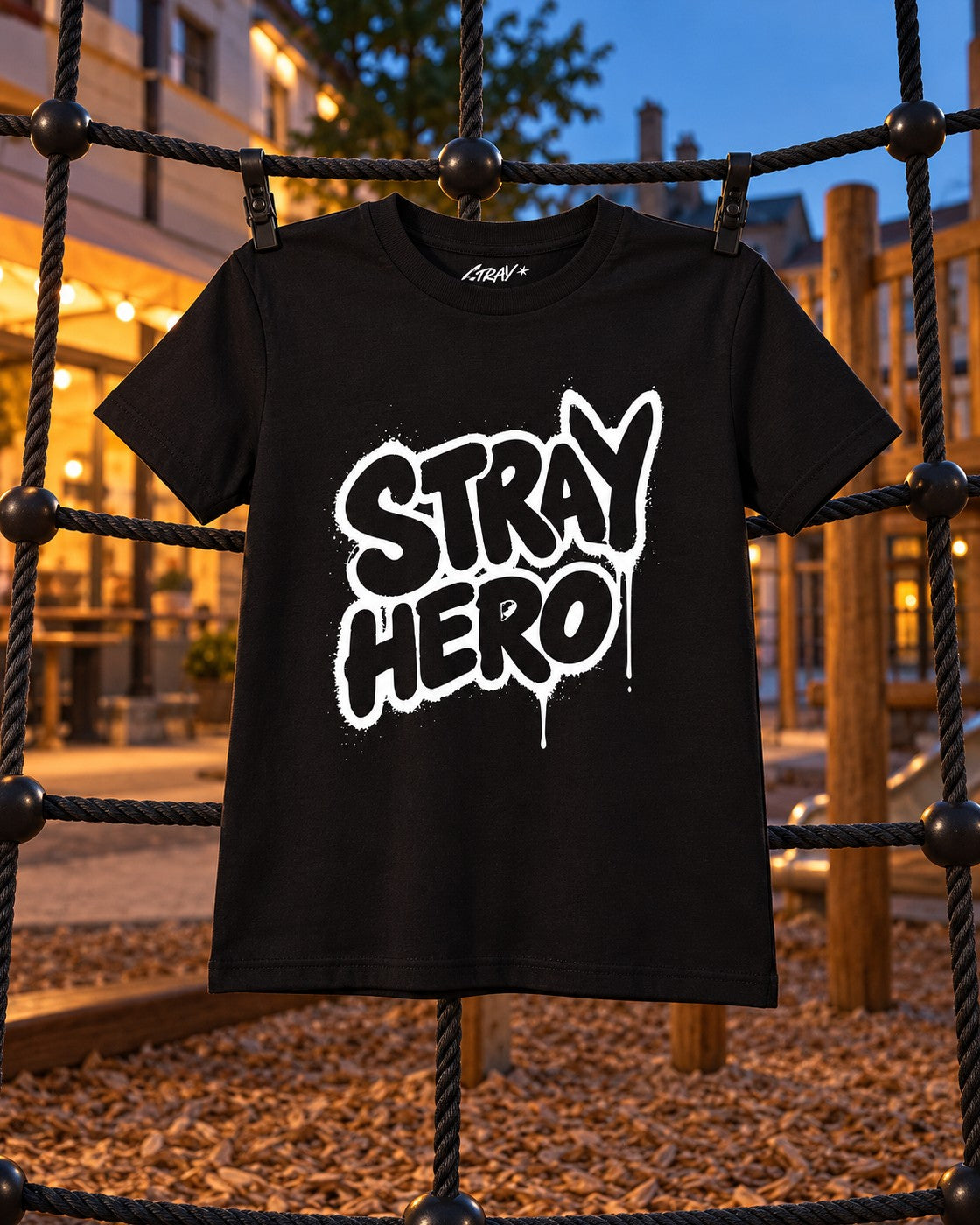 Stray Hero Kids Graffiti Tee, front print, black, clipped to a playground climbing net at dusk — AI-generated visual