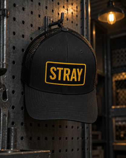 Tag Trucker Cap, front patch, black, hanging on a pegboard wall in a harbour workshop under a lamp — AI-generated visual
