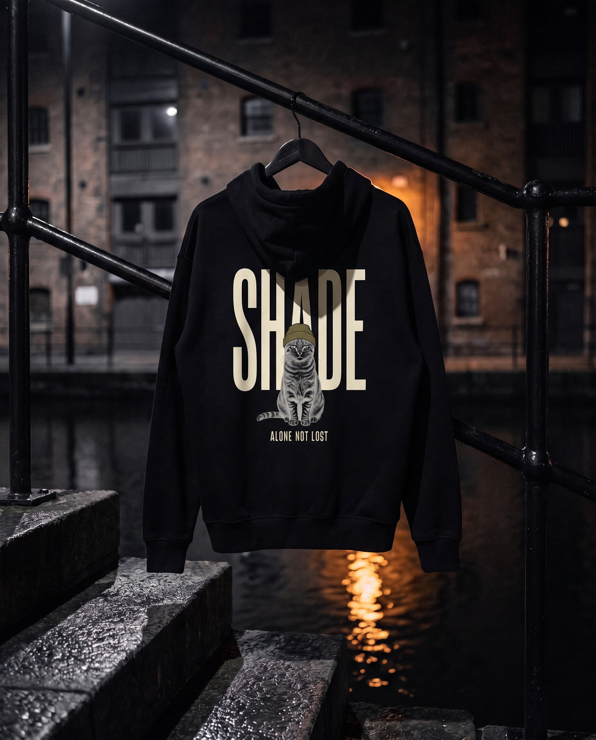 Unit 03 Shade Back Print Hoodie, back print, black, on a hanger at a canal-side railing at night — AI-generated visual