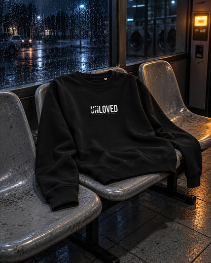 Unloved Sweatshirt, front print, black, draped over laundromat chairs by a rainy window at night — AI-generated visual