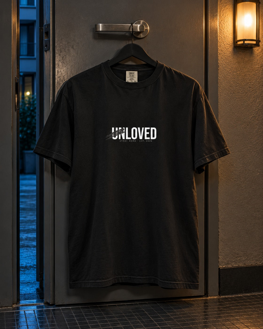 Unloved Tee, front print, black, hanging on a door handle under a wall lamp at night — AI-generated visual