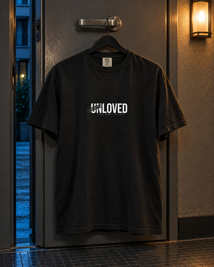 Unloved Tee, front print, black, hanging on a door handle under a wall lamp at night — AI-generated visual